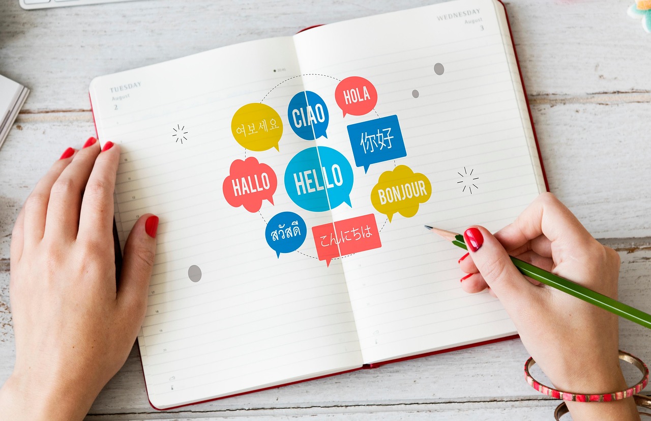 Student hands with red painted finger nails holding a pencil in right hand, ready to write in an notebook that has bubbles in which "hello" is written in various different languages