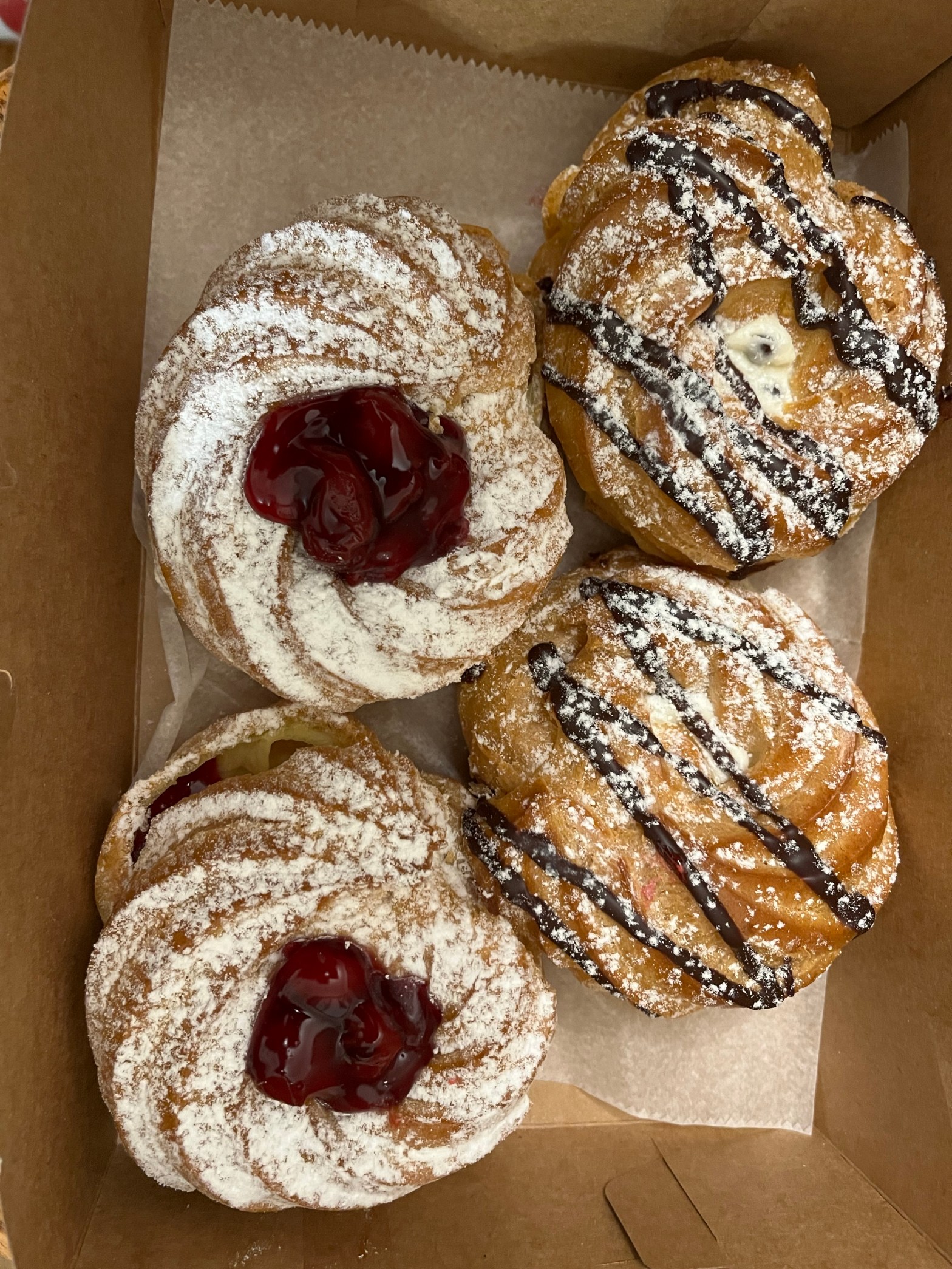 4 zeppole Italian pastries the two on the left are dusted with white powedered or confectioner's sugar and topped with sour cherries in syrup. The two on the left are dusted with powdered sugar and drizzled with chocolate